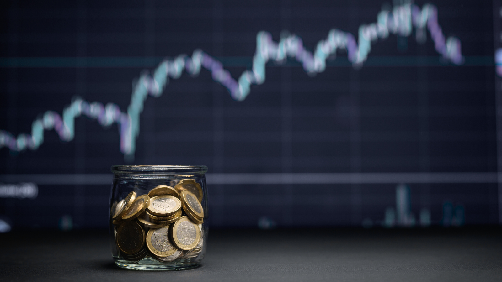Glass jar filled with coins in front of a blurred stock market chart, representing tech investing trends and capital growth.